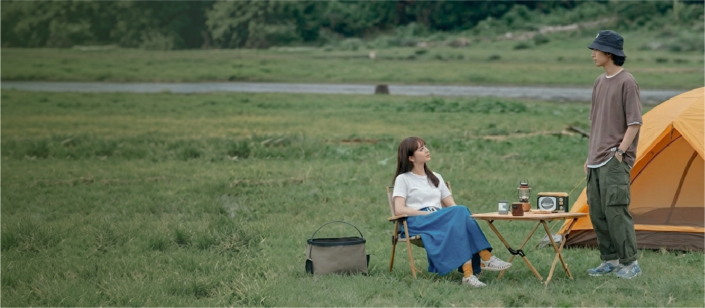 A wide outdoor lifestyle shot of three campers relaxing at a grassy campsite next to an orange tent. A woman in a mustard yellow bucket hat sits in a folding chair by a wooden table, while a man in a black Yama Workwear Bucket Hat stands nearby wearing an earth-toned outfit. The scene highlights rugged outdoor style and utility gear framed by an open field and natural greenery at Grandmama Store in Hong Kong.