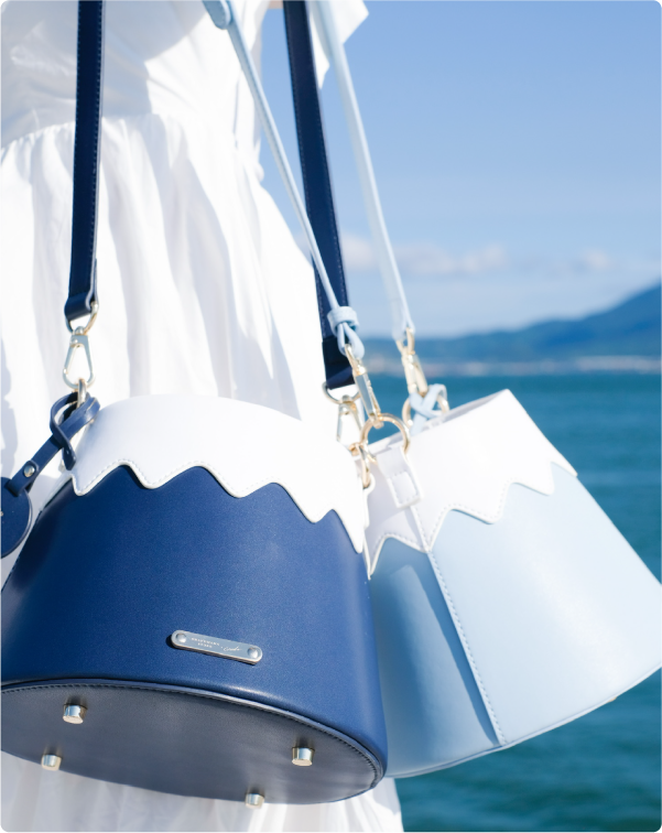 A close-up shot of a model in a white dress carrying two Mt. Fuji Goen Bags over her shoulder against an ocean background. The image highlights the contrast between the deep navy 'Night' bag in the foreground and the pastel 'Snow' blue bag beside it. It showcases their smooth leather texture, wavy snow-capped split-panel top, gold hardware clasps, and metal protective feet on the bottom at Grandmama Store in Hong Kong.