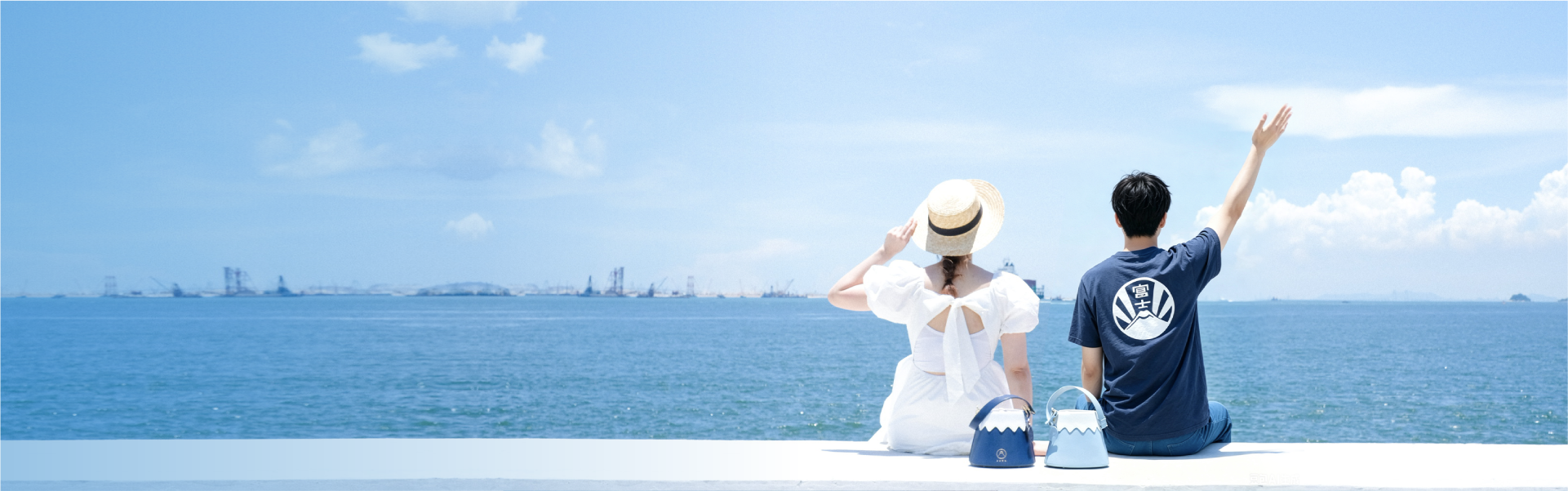 A wide seaside banner shot of a couple sitting on a pier under a bright blue sky. The woman on the left wears a white open-back dress paired with the French Straw Boater Hat, while the man on the right wears a navy Fuji graphic t-shirt. Placed between them are two Mt. Fuji Goen Bags in the 'Night' navy and 'Snow' light blue colourways, showcasing their wavy white split-panel designs from the Fuji Series at Grandmama Store in Hong Kong