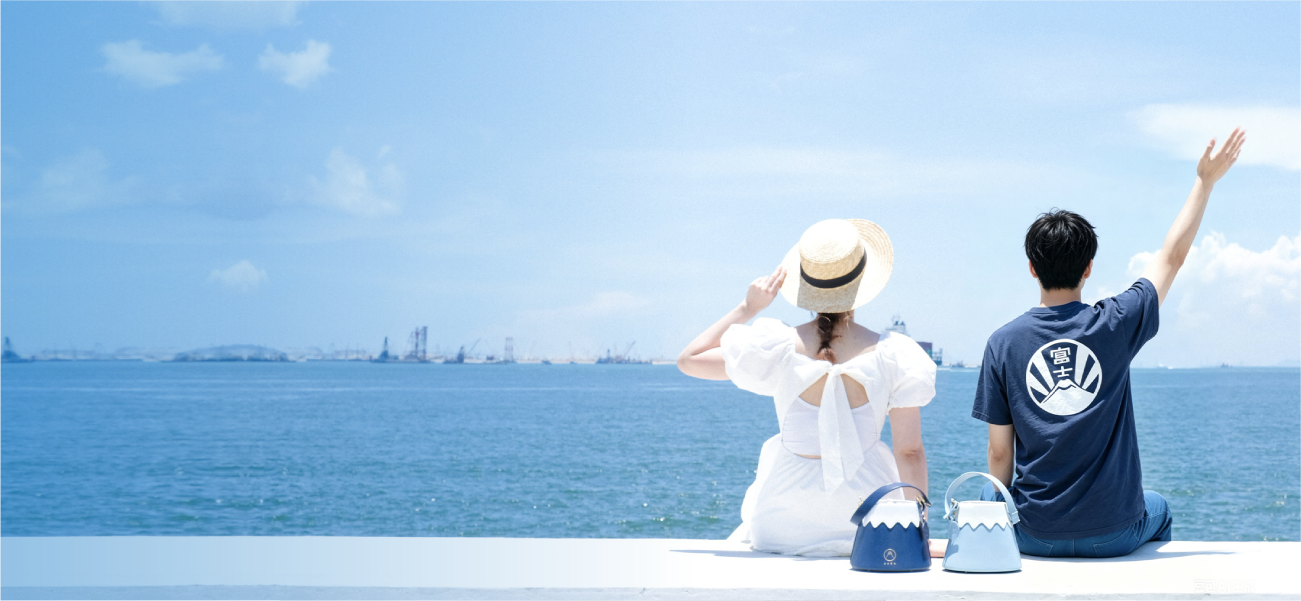 A wide seaside banner shot of a couple sitting on a pier under a bright blue sky. The woman on the left wears a white open-back dress paired with the French Straw Boater Hat, while the man on the right wears a navy Fuji graphic t-shirt. Placed between them are two Mt. Fuji Goen Bags in the 'Night' navy and 'Snow' light blue colourways, showcasing their wavy white split-panel designs from the Fuji Series at Grandmama Store in Hong Kong