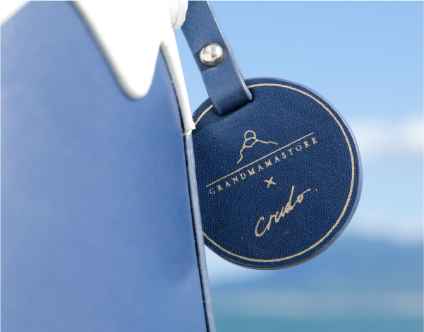 A detailed close-up shot focusing on the circular navy blue leather hangtag attached to the Mt. Fuji Goen Bag in the 'Night' colourway. The image highlights the fine leather grain, precise gold stitching around the rim, and gold-foil stamped text reading 'GRANDMAMA STORE X Crudo' with a mountain logo, set against a bright ocean sky background at Grandmama Store in Hong Kong.