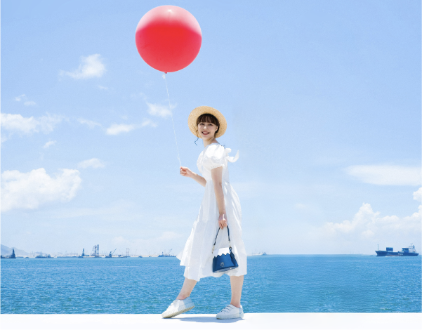 A full shot of a female model walking along a waterfront pier on a sunny day. She wears a white ruffled dress, sneakers, and the natural woven French Straw Boater Hat from the Fuji Series while holding a bright red balloon in one hand and carrying the Mt. Fuji Goen Bag in the navy 'Night' colourway in the other, framed by blue sea and sky at Grandmama Store in Hong Kong.