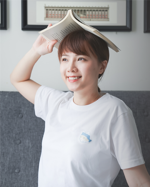 A playful portrait of a smiling woman holding an open book on her head while wearing the white Dancing Cat Tee with a small chest graphic at Grandmama Store in Hong Kong.