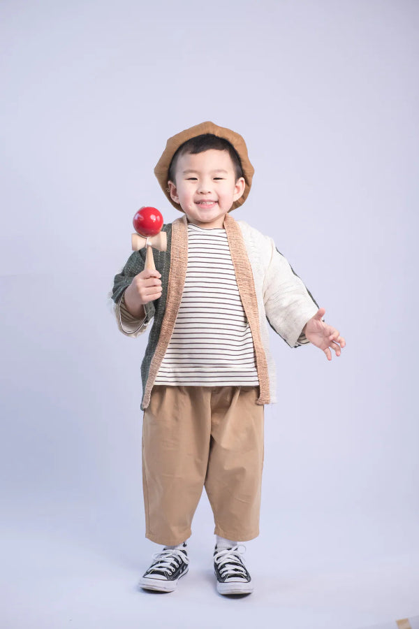 A young boy holding a kendama toy while wearing a Hand-cranked Kids Kimono jacket in the Yakushima olive green colorway at Grandmama Store in Hong Kong.