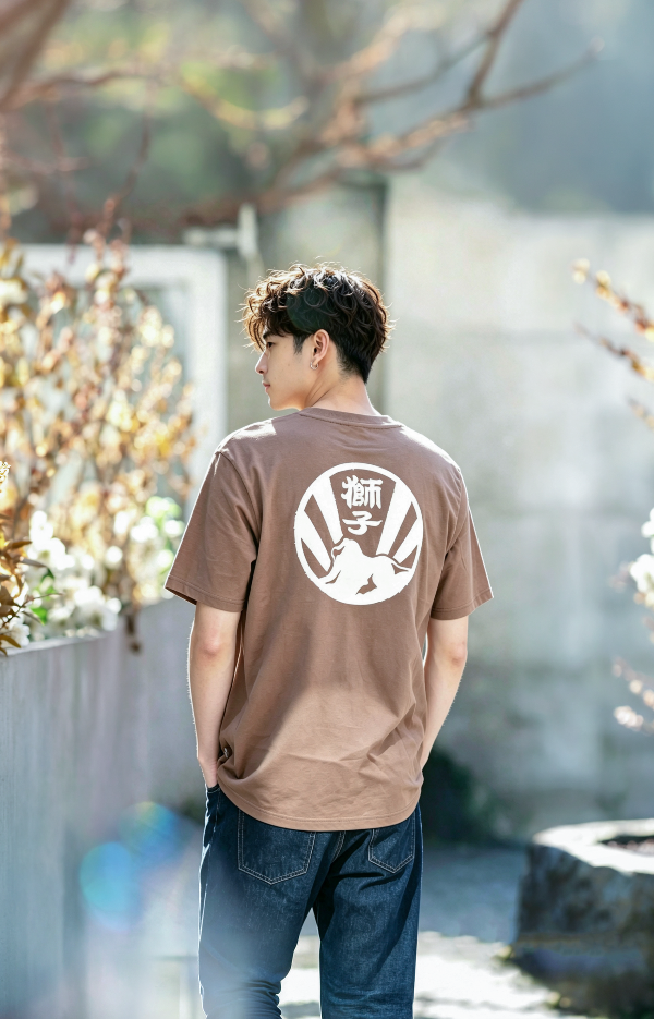 A young man outdoors wearing a brown Kamon Tee-LionRock t-shirt with a white circular back graphic at Grandmama Store in Hong Kong.