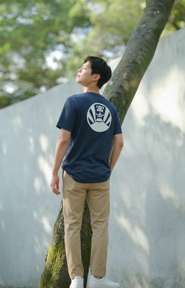 A young man standing outdoors in sunlight wearing a navy blue Kamon Tee-Fujisan t-shirt with a Mt. Fuji crest back graphic at Grandmama Store in Hong Kong.