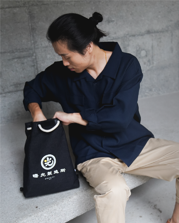 Seated male model accessing the Light of Dawn Sake Bag in Black Denim at Grandmama Store in Hong Kong.