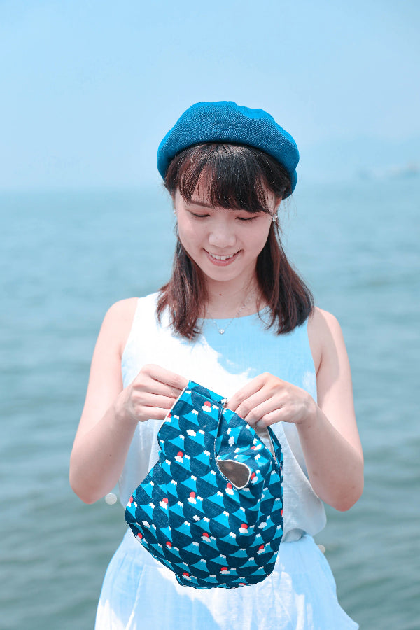 Smiling model opening a blue Fuji Handled Moonshell Bag outdoors by the sea at Grandmama Store in Hong Kong.