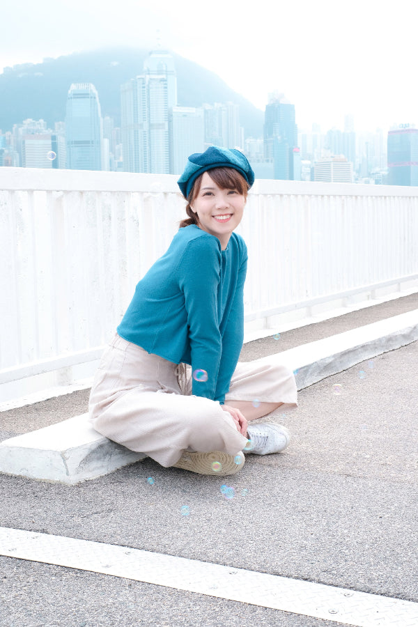 Smiling model sitting outdoors wearing a blue Sunshine Beret with city view at Grandmama Store in Hong Kong.