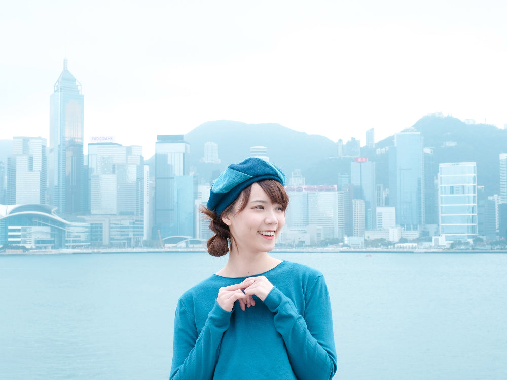Smiling woman wearing a blue Sunshine Beret with Hong Kong Victoria Harbour skyline background at Grandmama Store in Hong Kong.