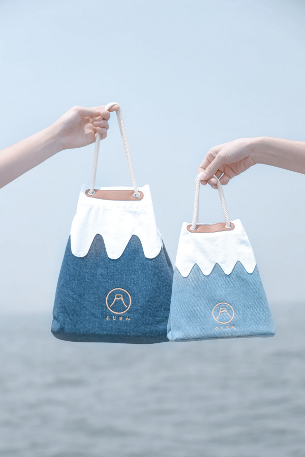 Two hands holding a medium navy blue and a small light blue Mt. Fuji sake bag against an ocean backdrop at Grandmama Store in Hong Kong.