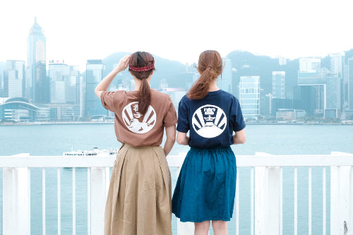 Two women standing at Victoria Harbour wearing Kamon Tee t-shirts in brown and navy blue showcasing their back prints at Grandmama Store in Hong Kong.