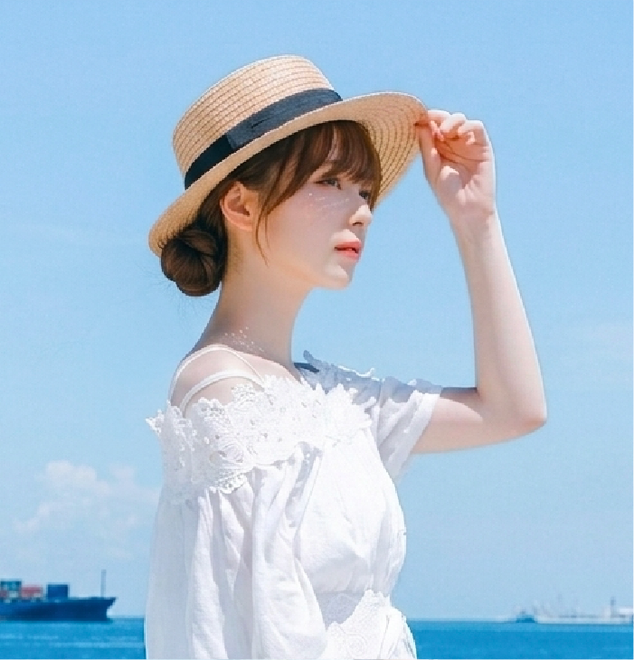 A side profile shot of a female model with a low bun hairstyle wearing the French Straw Boater Hat from the Fuji Series under a bright blue sky. She wears a white off-the-shoulder lace blouse and holds the brim of the natural woven straw hat, which features a classic flat crown, wide brim, and a dark black ribbon band. The shot highlights the fine straw braid texture against a coastal ocean backdrop at Grandmama Store in Hong Kong.